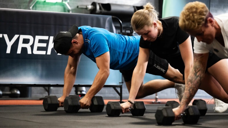 Drie sporters in een sportschool voeren de 'Load Crawl' oefening uit, waarbij ze in een lage kruiphouding naar voren bewegen en met hun handen steunen op zeshoekige dumbbells.