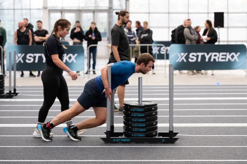 Een man in een blauw sportshirt en donkere korte broek leunt ver naar voren en duwt met grote inspanning een zware, met halterschijven beladen slee (sled push) over een grijze indoor atletiekbaan met witte lijnen. Een vrouwelijke scheidsrechter in zwarte sportkleding loopt aandachtig met hem mee. Op de achtergrond staan toeschouwers te kijken van achter blauwe Styrekx-spandoeken in een grote, lichte sporthal.