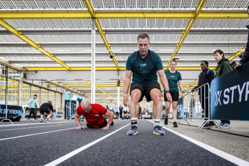 Twee mannelijke atleten voeren de 'Burpee Broad Jumps' oefening uit op een binnenbaan tijdens een fitnessevenement; de rechter atleet springt ver naar voren in de lucht, terwijl de linker atleet zich op de grond in de push-up fase bevindt.