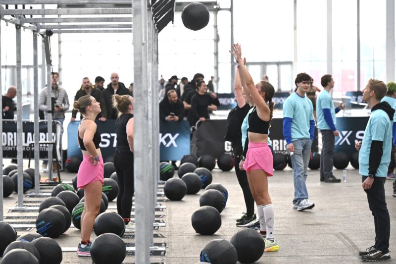 Een vrouwelijke atleet in een felroze broekje voert de 'Wall Ball' oefening uit door een grote zwarte medicijnbal hoog de lucht in te gooien naar een metalen rek, terwijl een andere deelneemster toekijkt.