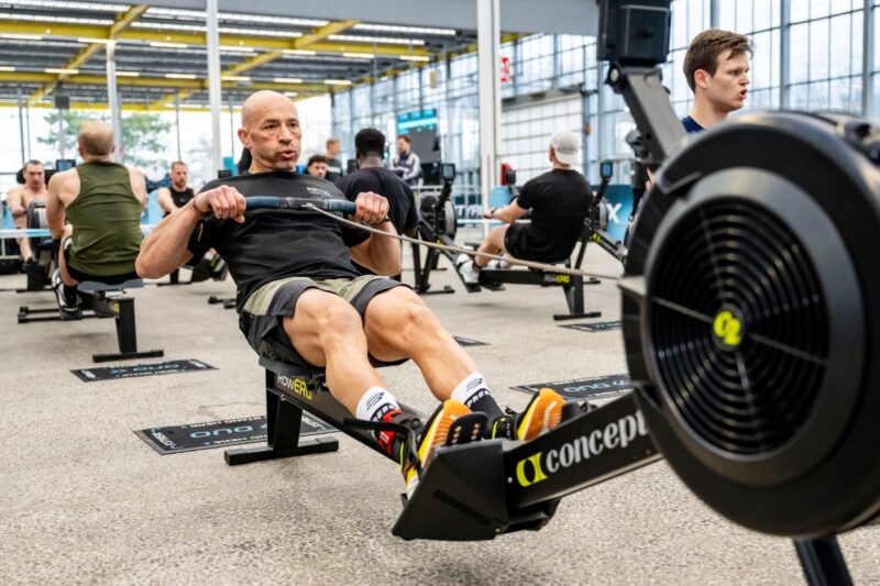 Een kale man in een zwart t-shirt en grijze sportbroek roeit fanatiek en geconcentreerd op een Concept2 RowErg roeitrainer. Op de achtergrond roeien meerdere andere mannen op hun eigen machines in een grote, lichte industriële sporthal tijdens een Styrekx evenement.