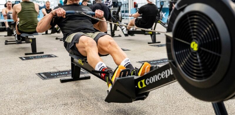 Een kale man in een zwart t-shirt en grijze sportbroek roeit fanatiek en geconcentreerd op een Concept2 RowErg roeitrainer. Op de achtergrond roeien meerdere andere mannen op hun eigen machines in een grote, lichte industriële sporthal tijdens een Styrekx evenement.