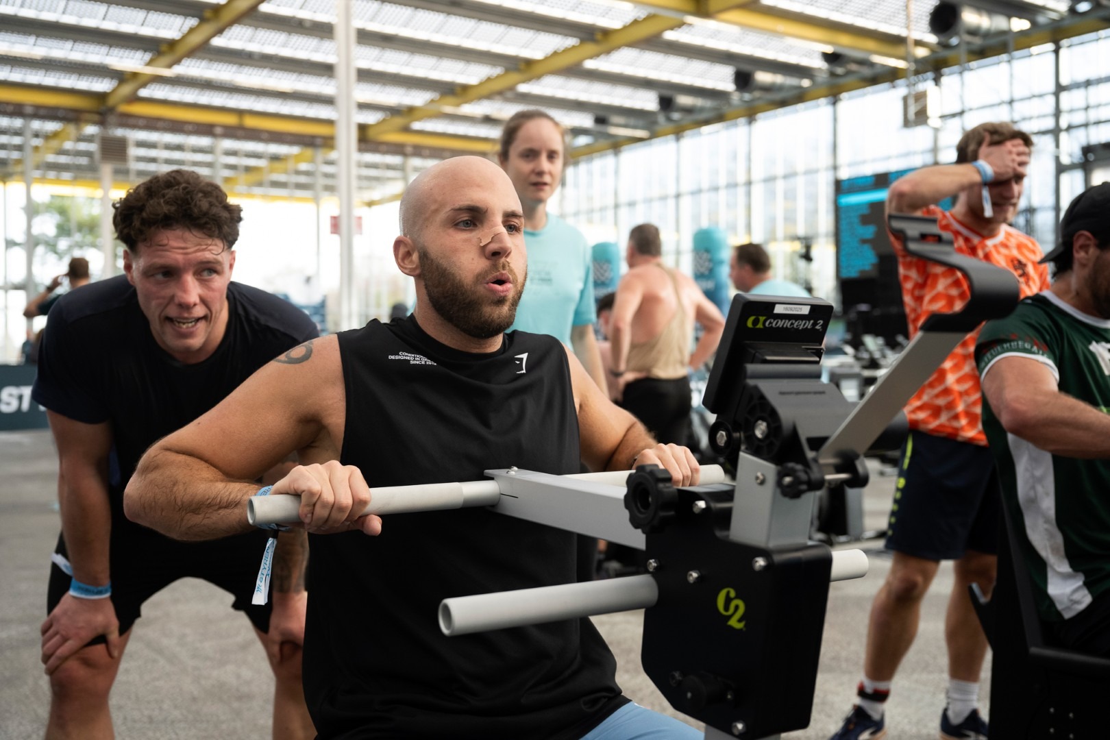 Een mannelijke atleet levert een zware inspanning tijdens de 'Strength Erg' oefening op een Concept2 apparaat, terwijl een medesporter of coach voorovergebogen naast hem staat om hem fanatiek aan te moedigen.