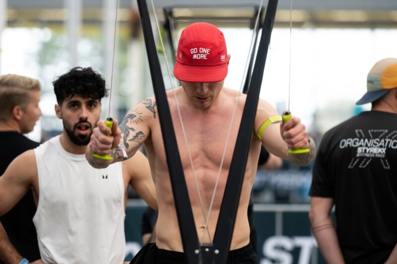 Een afgetrainde man met een ontbloot bovenlichaam, tatoeages op zijn arm en een rode pet met de tekst 'GO ONE MORE' trekt geconcentreerd en met veel inspanning aan de hendels van een SkiErg apparaat. Links achter hem kijkt een man in een wit shirt met een intense blik toe. Aan de rechterkant staat iemand op de rug gezien in een zwart shirt met de opdruk 'ORGANISATION STYREKX FITNESS RACE'.