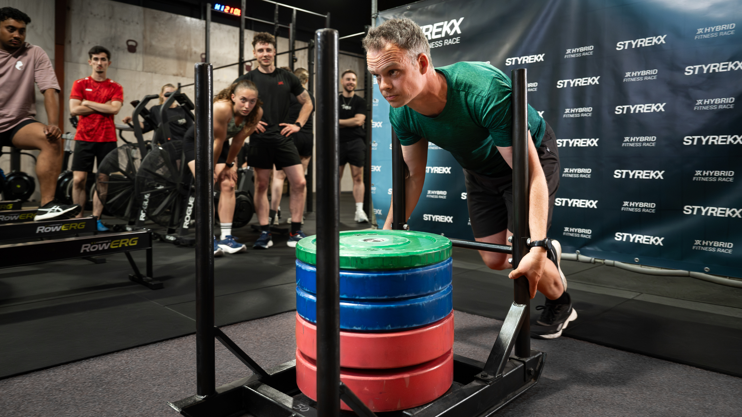 Man duwt zware sled tijdens STYREKX fitnessrace terwijl zijn team gefocust toekijkt in de arena.