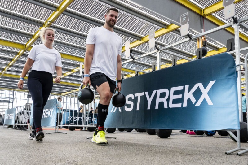 Een mannelijke sporter voert de 'Farmers Carry' oefening uit door met in elke hand een zware kettlebell naar voren te lopen, terwijl een vrouwelijke sporter naast hem loopt tijdens een fitnessevenement.
