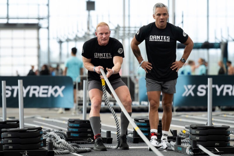 Een mannelijke atleet voert de 'Sled pull' oefening uit door met veel kracht aan een dik touw te trekken, terwijl een andere man met zijn handen in zijn zij staat toe te kijken tijdens een fitnesswedstrijd.